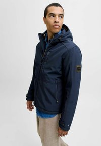 Veste imperméable bleu marine avec capuche, fermeture éclair à l'avant et poches latérales. Équipée de poignets ajustables et d'un patch logo sur la manche.