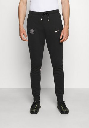 Mann trägt schwarze Nike-Sweathosen mit Paris Saint-Germain-Logo und schwarze Sportschuhe mit gelben Akzenten, steht auf grauem Boden.