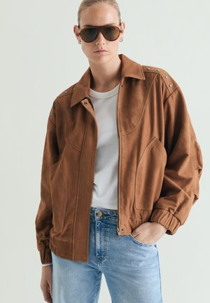 Personne portant une veste en daim marron avec des clous turquoise, un t-shirt blanc, un jean bleu et de grandes lunettes de soleil marron, debout avec une main dans la poche.