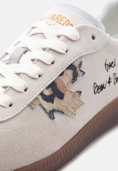 Baskets en suède beige avec lacets blancs, semelle en caoutchouc marron, et un patch en forme de feuille d'érable cousue avec des accents noirs et marron clair.