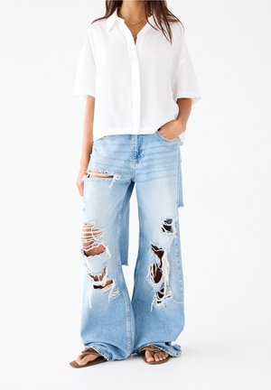 Kvinde iført løstsiddende, lyseblå meget slidte jeans, hvid kortærmet skjorte med knapper og sandaler, stående imod en hvid baggrund.