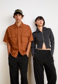 Chemise brune à manches courtes avec deux poches sur la poitrine portée par un homme, associée à un pantalon noir ; femme en veste noire cropped à fermeture éclair avec col montant et pantalon noir.