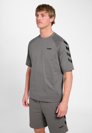 Mężczyzna w szarej koszulce hummel z krótkim rękawem i pasujących spodenkach z czarnymi paskami w jodełkę na prawej rękawie i lewej nodze.