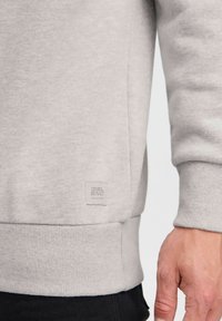 Sweatshirt gris clair en tissu doux, avec des poignets et un ourlet côtelés, et un petit label rectangulaire sur le côté inférieur.