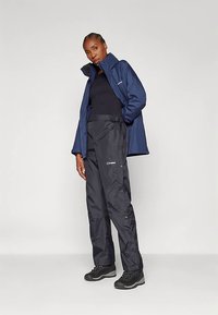 Γυναίκα με ναυτική ζακέτα, μαύρο τοπ, μαύρο αδιάβροχο παντελόνι και μαύρες ορειβατικές μπότες, στέκεται με το ένα χέρι στην τσέπη σε απλό φόντο.