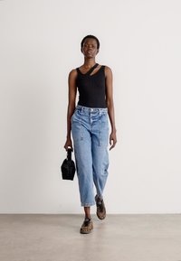 Top sin mangas negro con canalé, jeans de tiro alto azul claro y zapatillas con estampado. Sosteniendo un bolso pequeño negro, de pie contra un fondo liso.