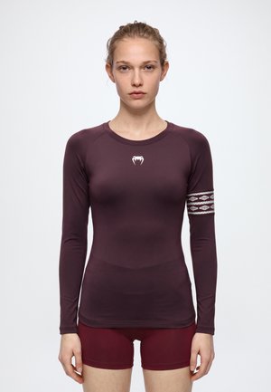 Femme portant un haut de sport à manches longues couleur prune, avec des bandes à motifs sur les bras et un short bordeaux, sur un fond blanc uni.