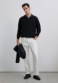 Camisa polo negra de manga larga, pantalones rectos blancos, chaqueta de cuero negra colgada del brazo y zapatos negros. Fondo neutro.