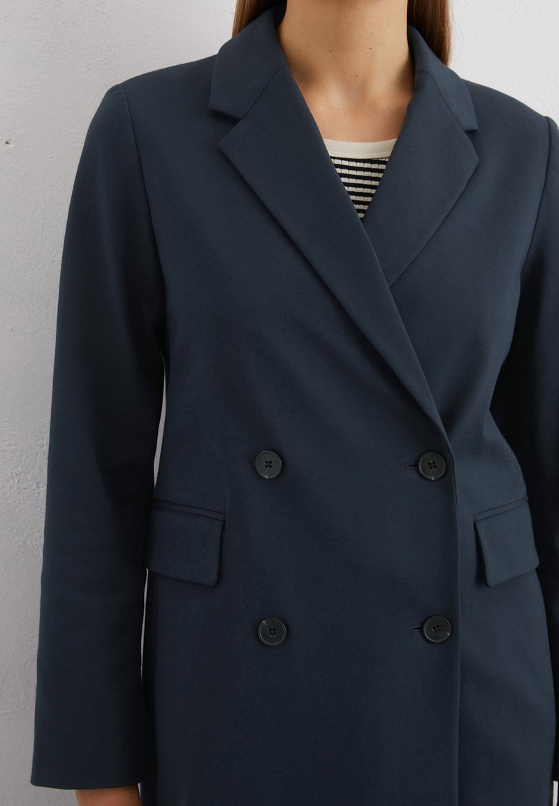 Marineblauer, zweireihiger Mantel mit Reverskragen, zwei Fronttaschen und schwarzen Knöpfen. Glattes Gewebe mit strukturierter Passform.