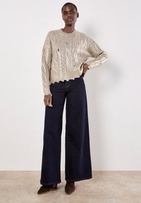 Maglione lavorato a maglia beige con motivo a treccia testurizzato, vestibilità oversize e polsini a costine, abbinato a jeans in denim scuri a gamba larga e stivali con tacco marroni.