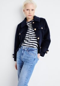 Chaqueta de mezcla de lana de color marine con botones dorados, combinada con una camisa a rayas en azul marino y blanco y jeans de color azul claro. Textura suave, diseño clásico.