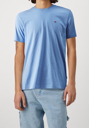 Homme portant un t-shirt à col rond bleu clair avec un petit logo de drapeau norvégien sur la poitrine et un jean en denim bleu clair.
