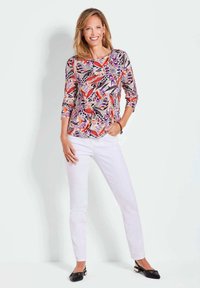 Bunt gemustertes Oberteil mit lebendigen roten, lila und schwarzen Designs; kombiniert mit figurbetonten weißen Hosen und schwarzen Ballerinas.