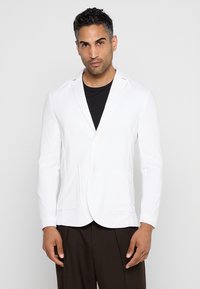 Blazer bianco con chiusura a bottoni, vestibilità slim, due tasche frontali e colletto a revers, abbinato a una T-shirt nera e pantaloni marroni.