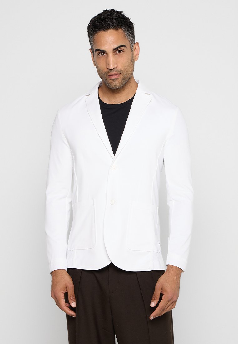 Blazer bianco con chiusura a bottoni, vestibilità slim, due tasche frontali e colletto a revers, abbinato a una T-shirt nera e pantaloni marroni.