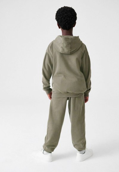 Oliven grøn hætte sweatshirt og matchende sweatpants lavet af blødt stof, med en afslappet pasform og ribbede manchetter. Hvide sneakers fuldender outfittet.