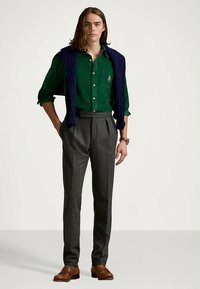 Mlad moški z dolgimi lasmi, oblečen v zeleno srajco, mornarsko pulover, položenega čez ramena, sive nagubane hlače in rjave usnjene čevlje.