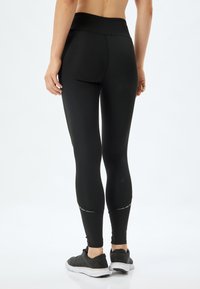 Zwarte legging met een hoge tailleband, gladde textuur en reflecterende accenten nabij de enkels. Gepaard met zwarte sportschoenen.