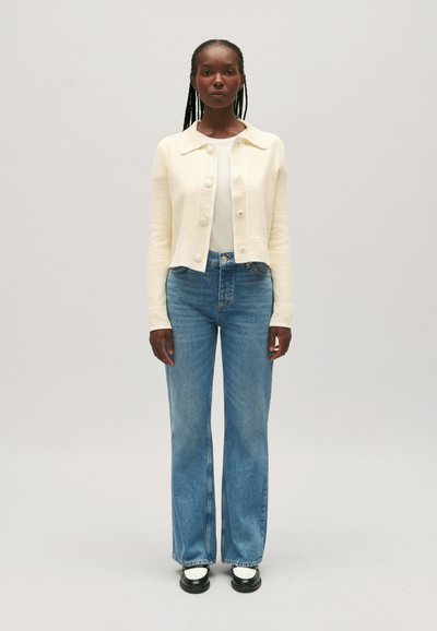 Cárdigan texturizado en crema con botones en la parte frontal, usado sobre una blusa blanca, combinado con jeans de campana azul claro y zapatos de plataforma negros.