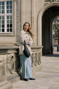 Femme en manteau beige et jean bleu clair appuyée sur une balustrade en pierre ornée devant un bâtiment historique avec une entrée en arc.