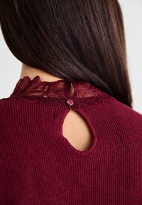 Bordeaux geribbelde top met kantafwerking bij de kraag, met een sleutelgatoopening aan de achterkant die is beveiligd met een decoratieve knop.