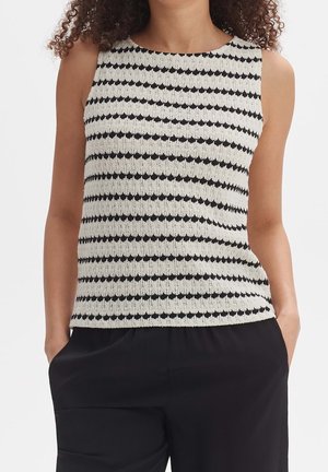 Vrouw in een mouwloze zwart-wit gestreepte gebreide top, gecombineerd met losse zwarte broek, handen in de zakken, tegen een effen achtergrond.