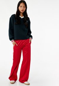 Felpa nera con scollo a V e profilo bianco, abbinata a pantaloni rossi a gamba larga con strisce bianche sui lati e una texture liscia.