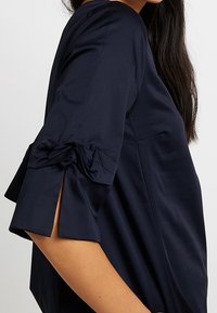 Mörkblå blus med korta ärmar, dekorerad med band och klippta manschetter, tillverkad av ett mjukt tyg med en avslappnad passform.