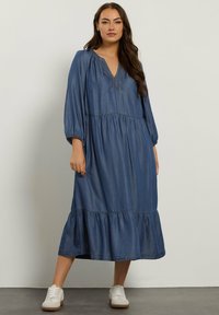 Denimmekko, jossa on V-pääntie, pitkät puff-hihat ja kerroksellinen helma. Sininen väri hienovaraisilla ompeluyksityiskohdilla. Yhdistetty valkoisiin tennareihin.