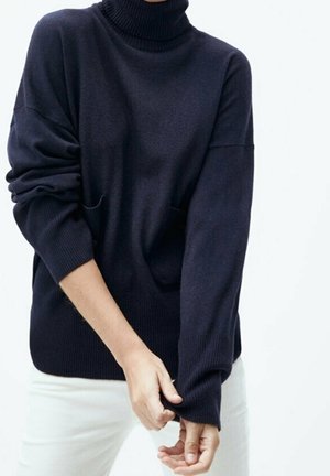 Pull-over col roulé marine à manches longues, col côtelé, coupe ample et poches latérales, porté avec un pantalon de couleur claire. Texture douce.
