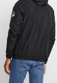 Sort vandtæt jakke med hætte, elastisk talje og manchetter. Har et rundt logomærke på venstre ærme. Bruges sammen med blå jeans.