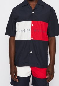 Camicia a maniche corte con blocchi di colore blu navy, bianco e rosso; presenta la scritta "HILFIGER" in blu e bottoni sul davanti.