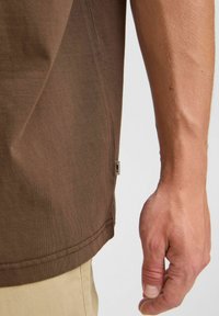 Camiseta de color marrón oscuro con una textura acanalada, que presenta una etiqueta en la costura lateral. La camiseta tiene un corte holgado y se combina con pantalones beige.