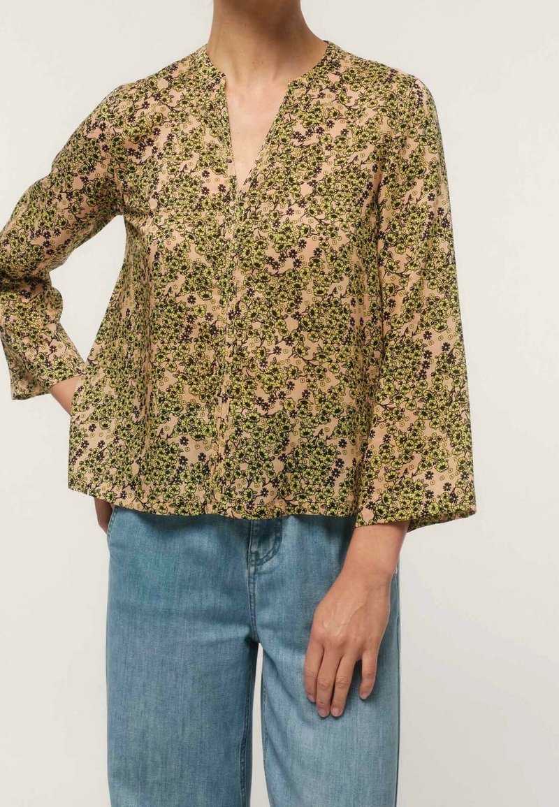 Blouse imprimé floral en beige avec des motifs verts et noirs, dotée d'un col en V, de manches trois-quarts et d'une coupe décontractée. Portée avec un jean bleu clair.