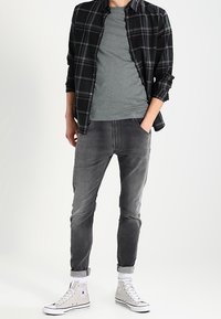Man som bär en svart och grå rutig skjorta över en grå t-shirt, slim-fit gråa jeans som är uppvikta i fållarna, och vita högsneakers, står med ena handen i fickan.