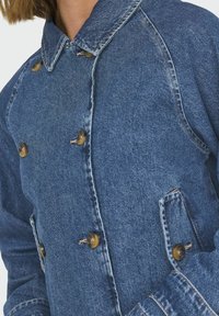 Džínová bunda s předním zapínáním na knoflíky, límcem a dvěma bočními kapsami. Má hnědé knoflíky a klasické modré praní s viditelným šitím.