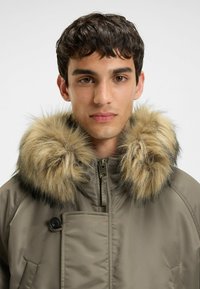 Khaki parka jacket met een nepbontkraag in beige en donkere tinten, voorzien van een ritssluiting en een knoopdetail op de borst.