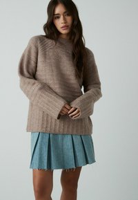 Beżowy sweter z dzianiny o fakturowanym wzorze i ściągaczami, zestawiony z jasnoniebieską spódnicą jeansową z plisami i postrzępionym dołem.