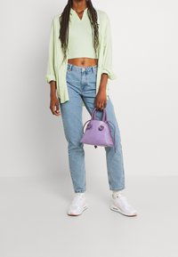 Top court vert clair, chemise oversized vert clair, jeans bleus et baskets blanches avec des accents roses ; tenant un petit sac violet texturé.