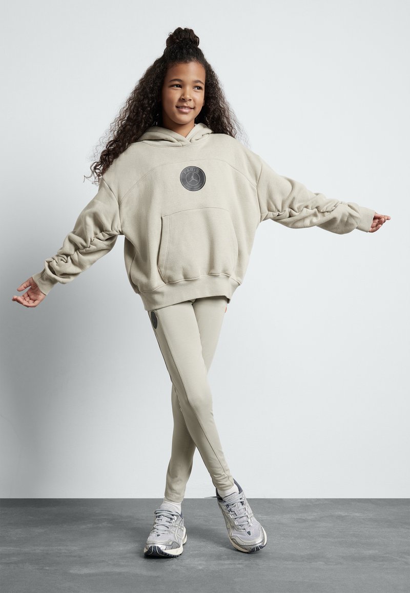 Felpa oversize beige con tasca frontale e patch con logo, abbinata a leggings aderenti coordinati; indossata con scarpe da ginnastica grigie chiare.
