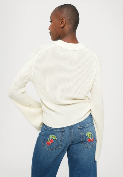 Persoon in een witte geribbelde trui en blauwe jeans met geborduurde rode kersen op de achterzakken, met de rug naar de camera gekeerd tegen een effen achtergrond.