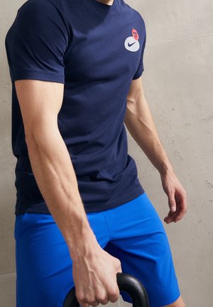 Ein Mann mit marineblauem Hemd und blauen Shorts hält eine Kettlebell mit einer Hand vor einem schlichten hellen Wandhintergrund.