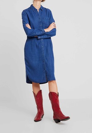 Kvinde iført en blå denim kjole med knapper og lange ærmer samt røde cowboystøvler, stående mod en ensartet lys grå baggrund.