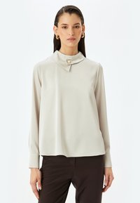 Blusa de un beige claro con cuello doblado y detalles de botones, mangas largas y una textura lisa, combinada con pantalones marrón oscuro.