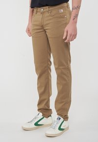 Chino beige con una vestibilità slim, caratterizzati da un design cucito, tasche frontali e una piccola etichetta con logo. Abbinati a sneakers bianche con dettagli verdi.