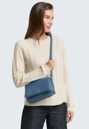 Helle beigefarbene Rippstrickpullover mit langen Ärmeln, kombiniert mit einer kleinen blauen Ledertaschen über der Schulter, die rechteckig geformt und mit einem verstellbaren Gürtel ausgestattet ist.