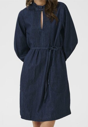 Femme portant une robe en jean foncé à manches longues, col montant, plis sur le devant, poches avant et ceinture nouée à la taille.