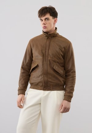 Joven con una chaqueta de cuero marrón con bolsillos de solapa y pantalones blancos, de pie frente a un fondo gris claro liso.