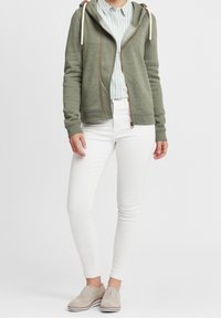 Sudadera de color verde oliva con cremallera y cordones, combinada con una camiseta de rayas azul claro, pantalones blancos ajustados y zapatos beige con cordones.
