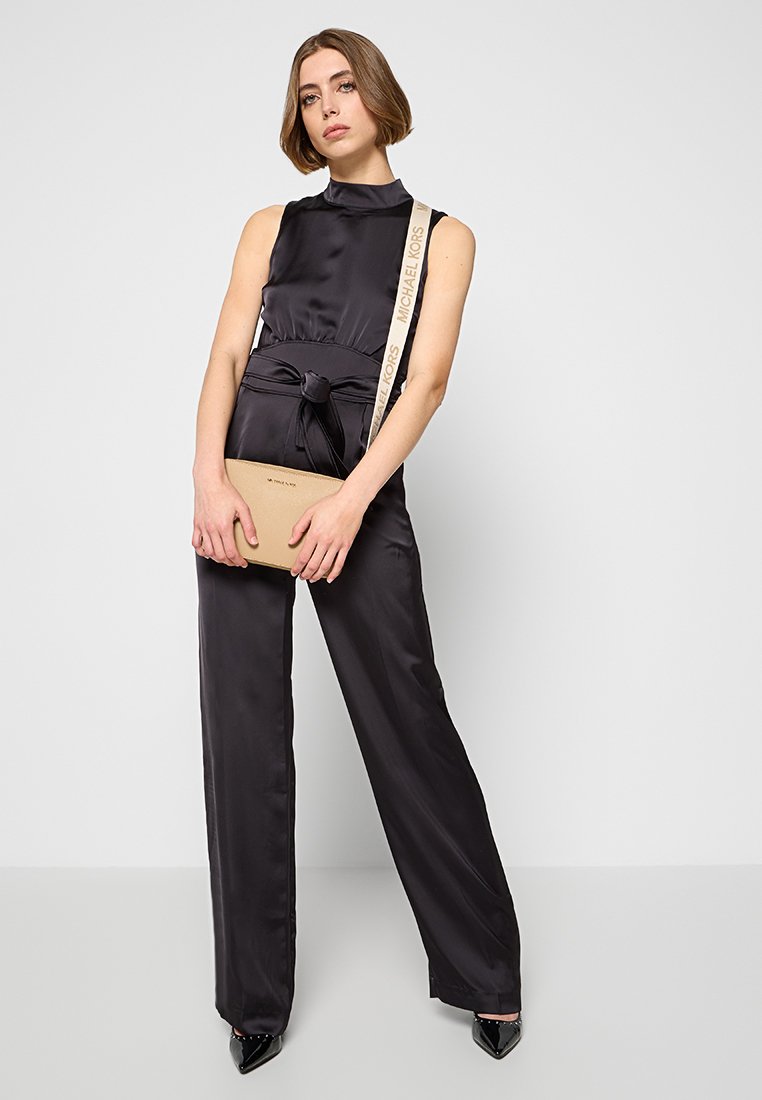 Combinaison en satin noir avec un col haut et une taille ceinturée. Modèle tenant un sac bandoulière beige avec une sangle "Michael Kors" et des talons aiguilles.